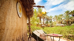 A rustic outdoor seating area with a woven chair and a table, surrounded by lush greenery and palm trees. - Riad Lamane (Zagora)