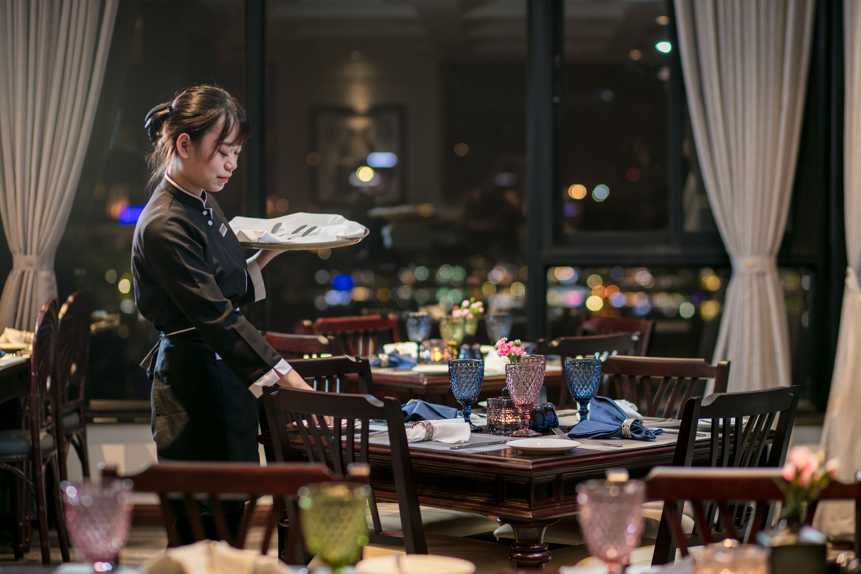 A restaurant setting with a waiter serving a dish. Tables are set with blue glassware, and there are floral centerpieces on the tables.