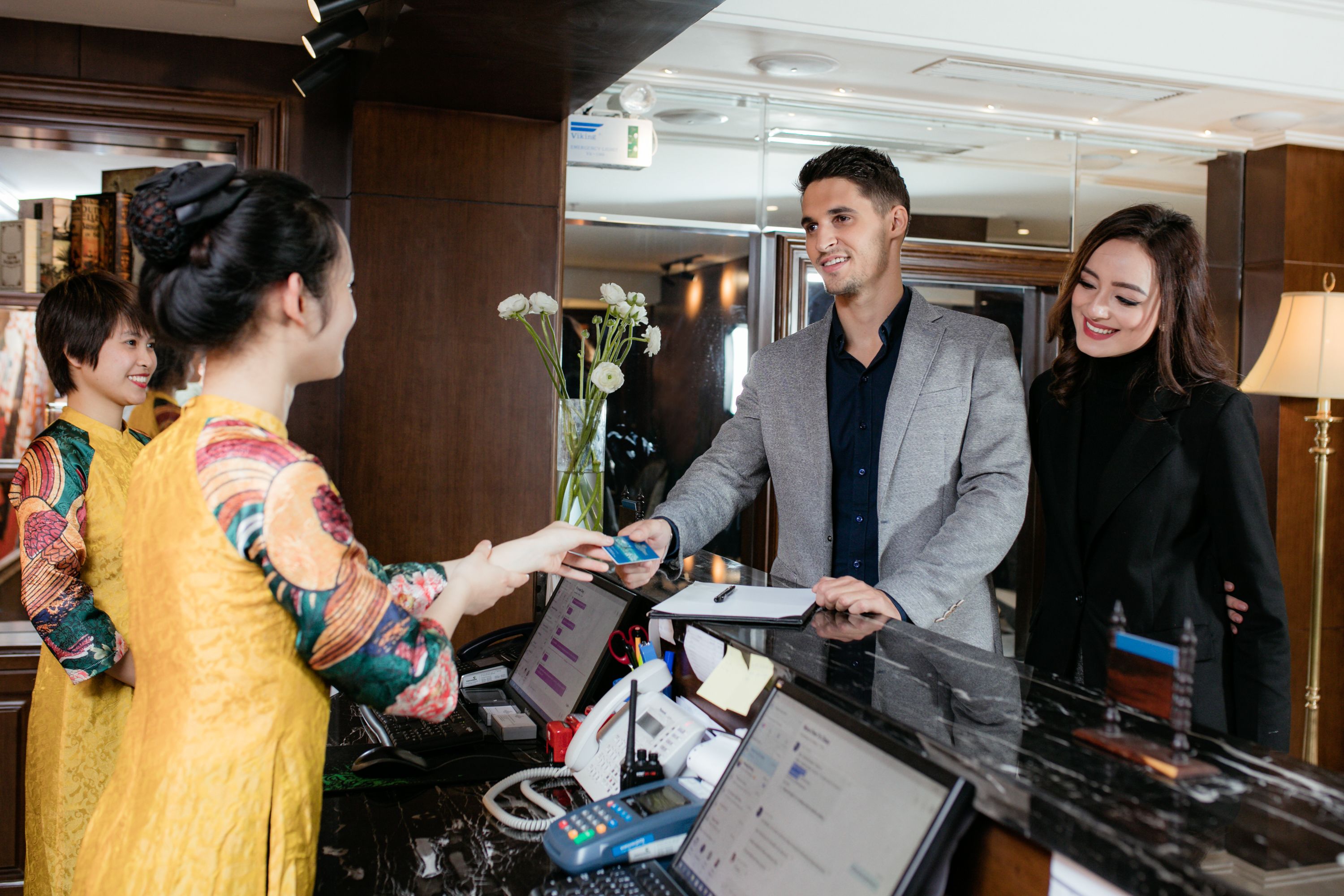 A hotel reception desk with a computer, phone, and a vase of flowers.