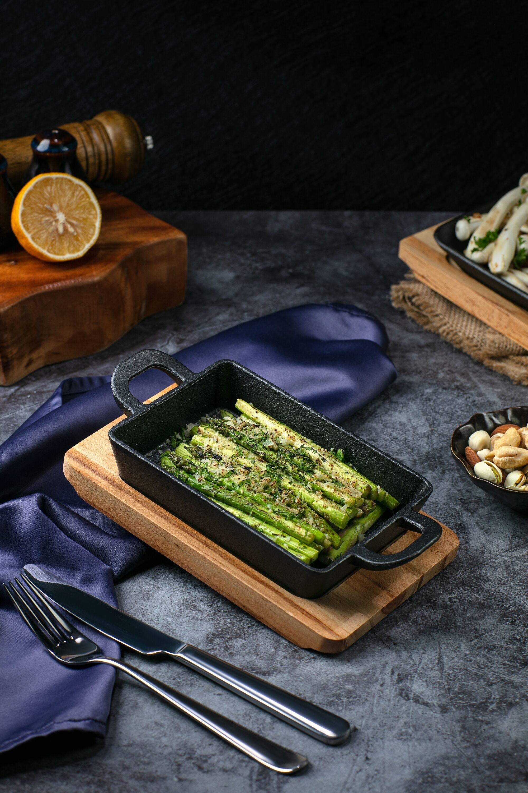 A cast iron skillet with grilled asparagus on a wooden board, accompanied by a fork and knife on a dark cloth.