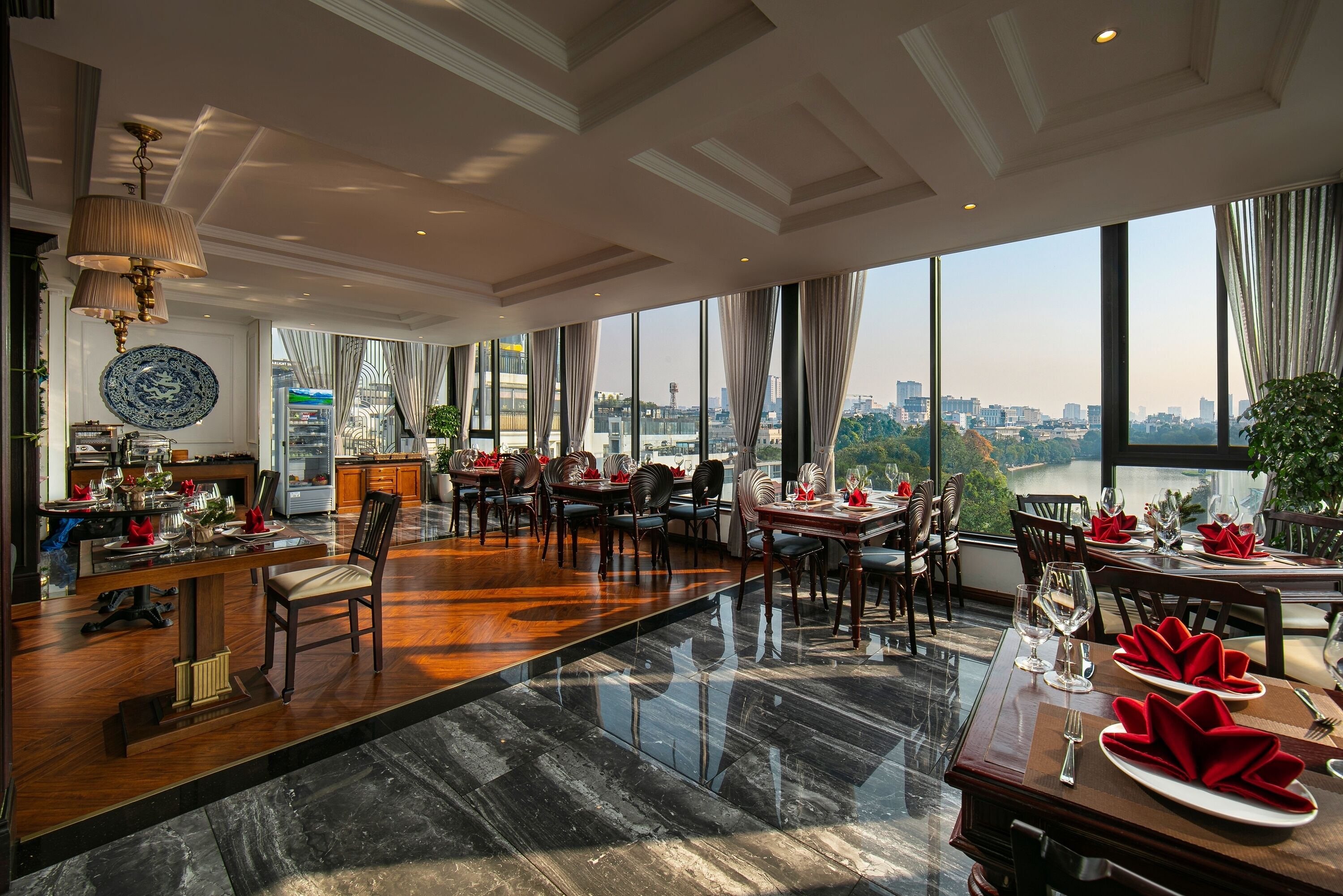 A spacious dining area with a high ceiling, large windows offering a city view, and a polished marble floor.