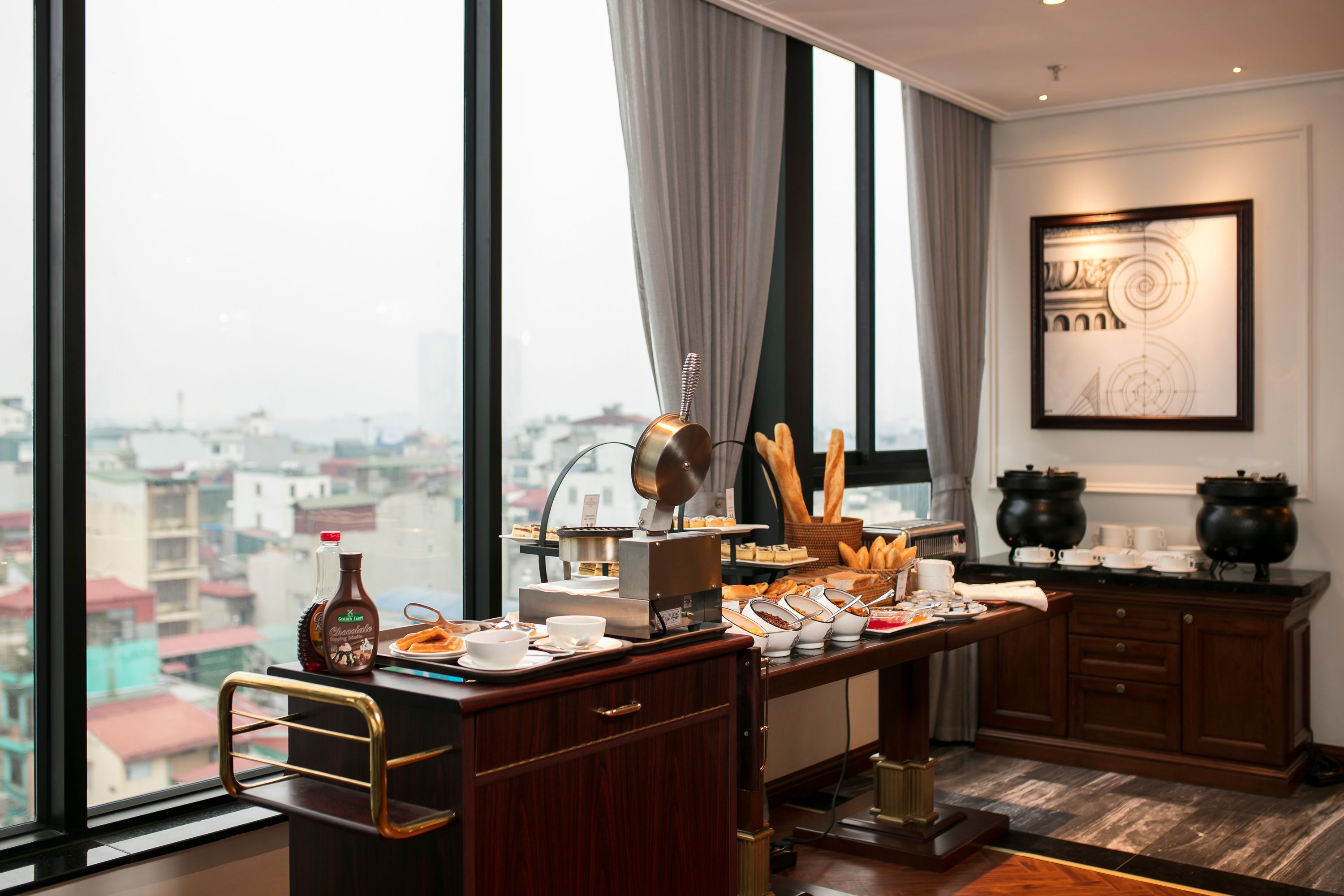 A modern hotel room with a large window offering a city view, a desk with breakfast items, and a decorative wall piece.