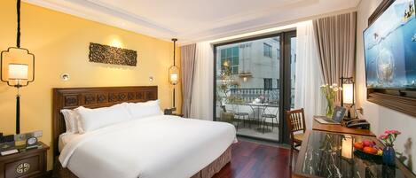 A hotel room with a large bed, a desk with a laptop, a vase with flowers, and a view of the city through the window.