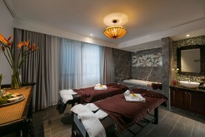 A spa room with two massage tables, a large window with curtains, a decorative wall, a sink, and a mirror.