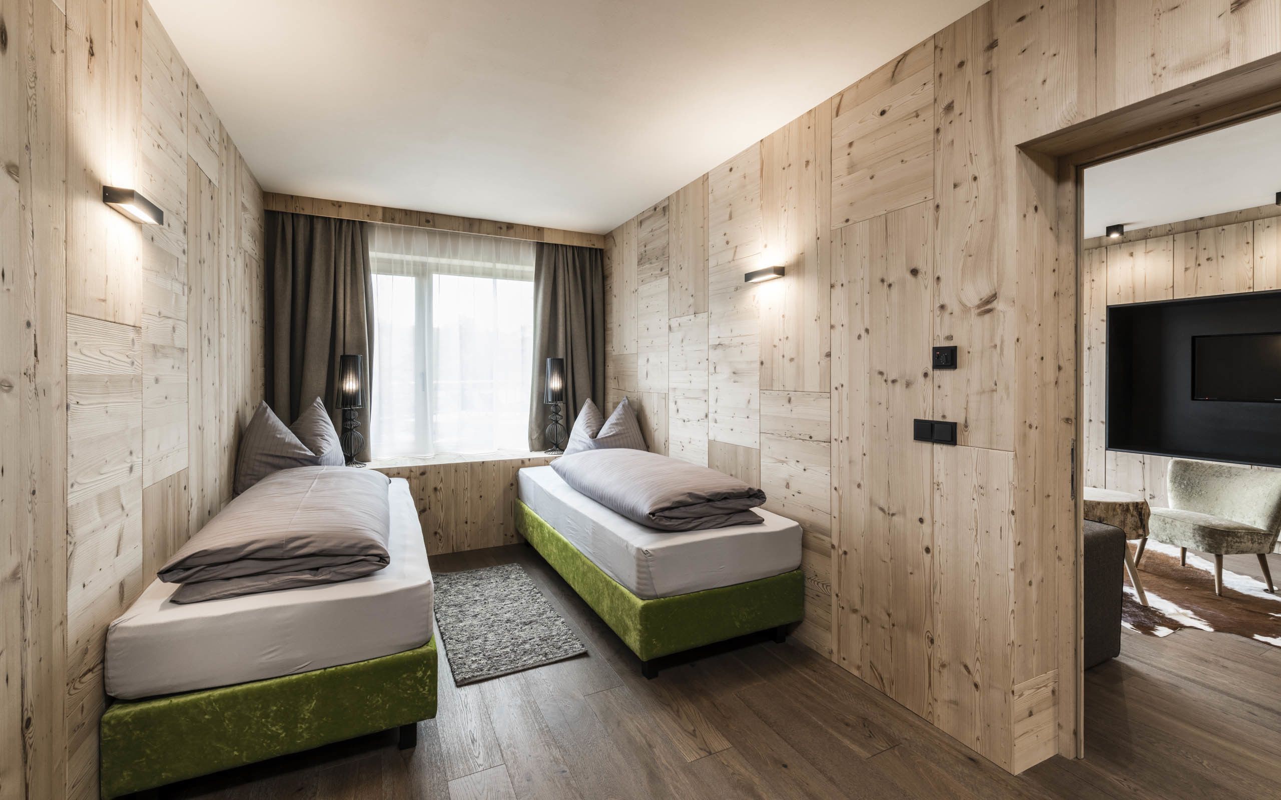 A room with two beds, wooden walls, a window with curtains, a TV mounted on the wall, and a small seating area with a chair and a table.