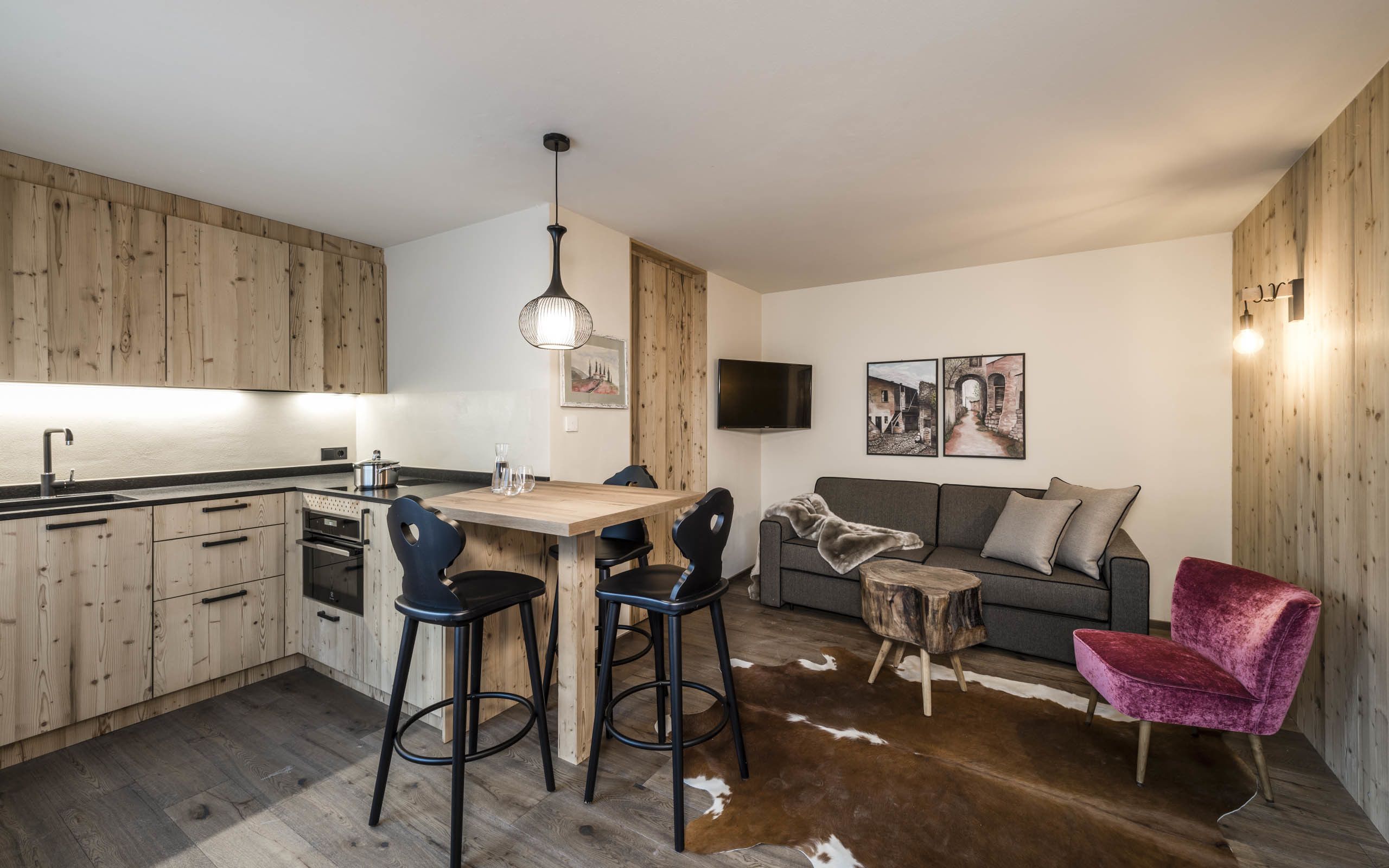 Una cocina moderna con armarios de madera, un comedor con taburetes, un sofá gris, una silla roja y una pequeña mesa de madera.