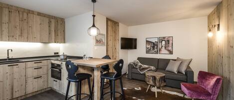 Una cocina moderna con armarios de madera, un comedor con taburetes, un sofá gris, una silla roja y una pequeña mesa de madera.
