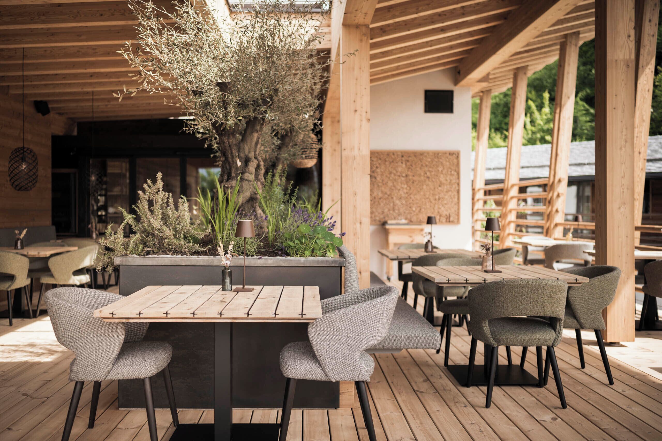 Een buitenterras met houten tafels en stoelen, omringd door planten en een houten pergola.