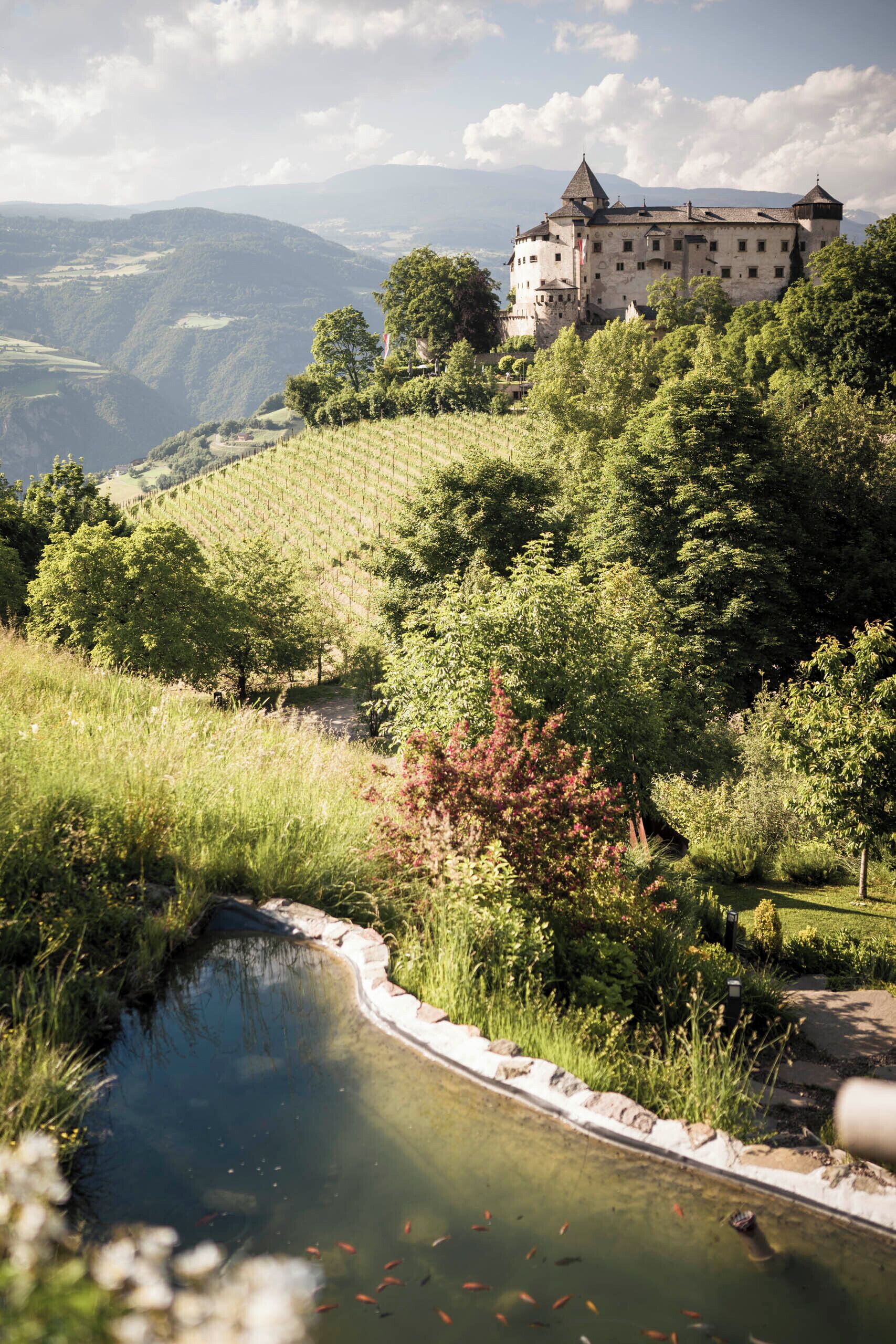 A castle on a hill with a pond and surrounding greenery.