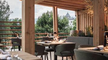 Un espace de restauration en plein air agrémenté de poutres, de tables et de chaises en bois, offrant une vue panoramique sur un paysage verdoyant, avec des arbres et un bâtiment visible au loin.