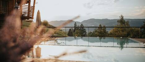 Una piscina infinita con terraza de madera, una estructura de madera con balcón y vistas a montañas distantes.