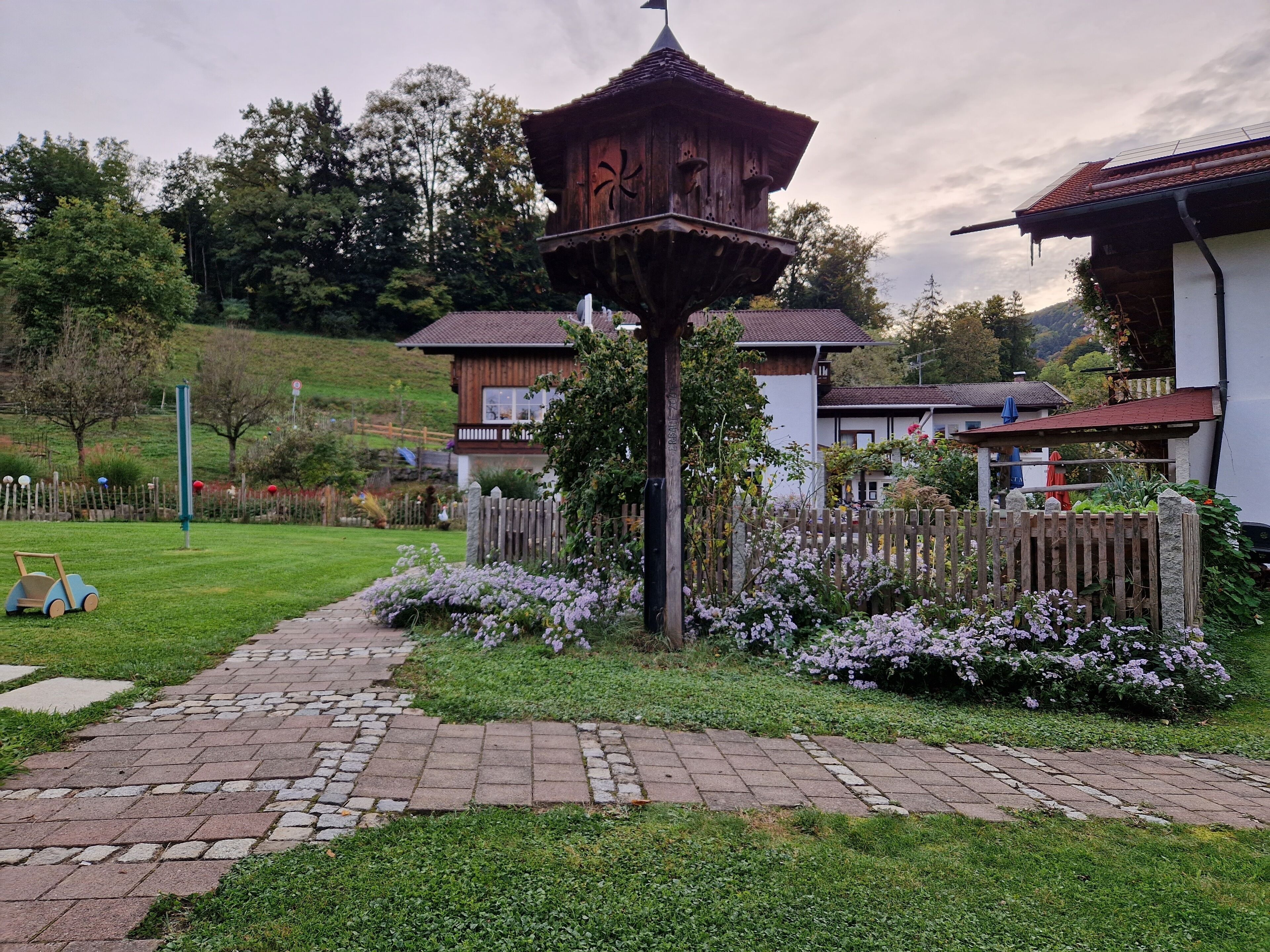 A traditional wooden gazebo in a garden with a paved walkway, surrounded by flowering plants and a wooden fence.