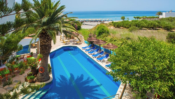 Ein Swimmingpool mit Liegestühlen und Sonnenschirmen, umgeben von Palmen und Blick auf den Strand.