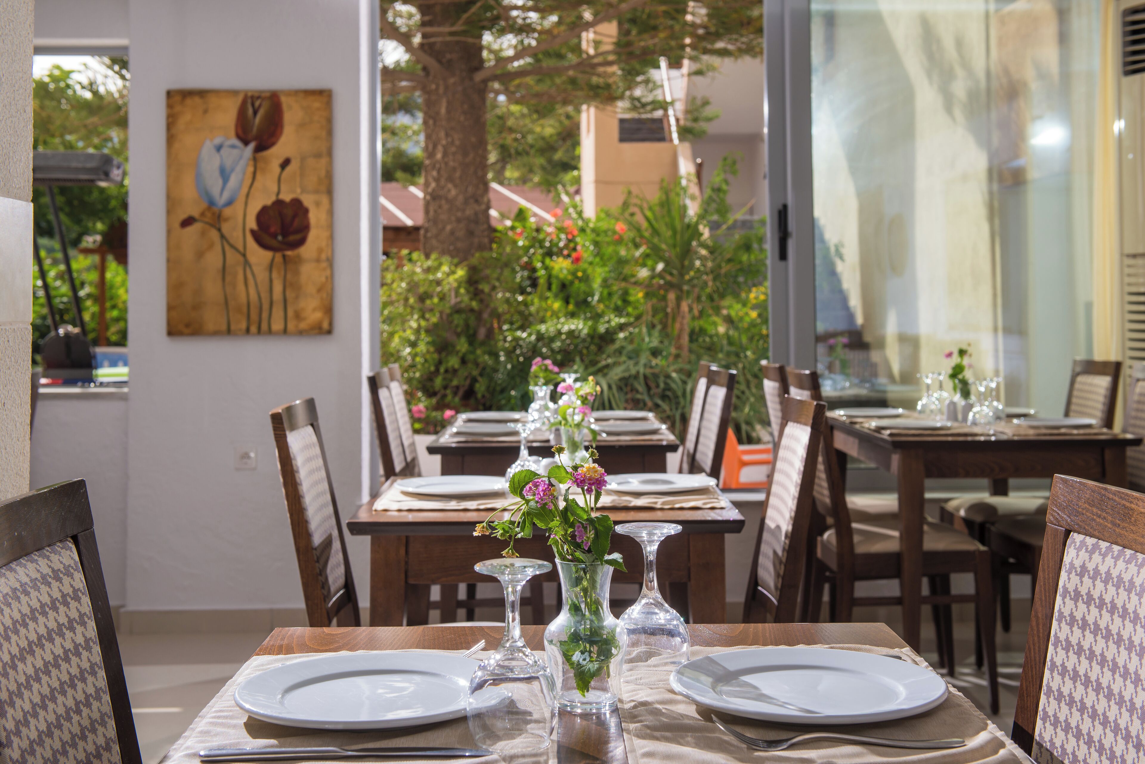 A dining area with tables set for two, each with a white plate, a glass, and a vase with flowers. There is a painting on the wall and a view of greenery outside.