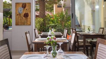 A dining area with tables set for two, each with a white plate, a glass, and a vase with flowers. There is a painting on the wall and a view of greenery outside.