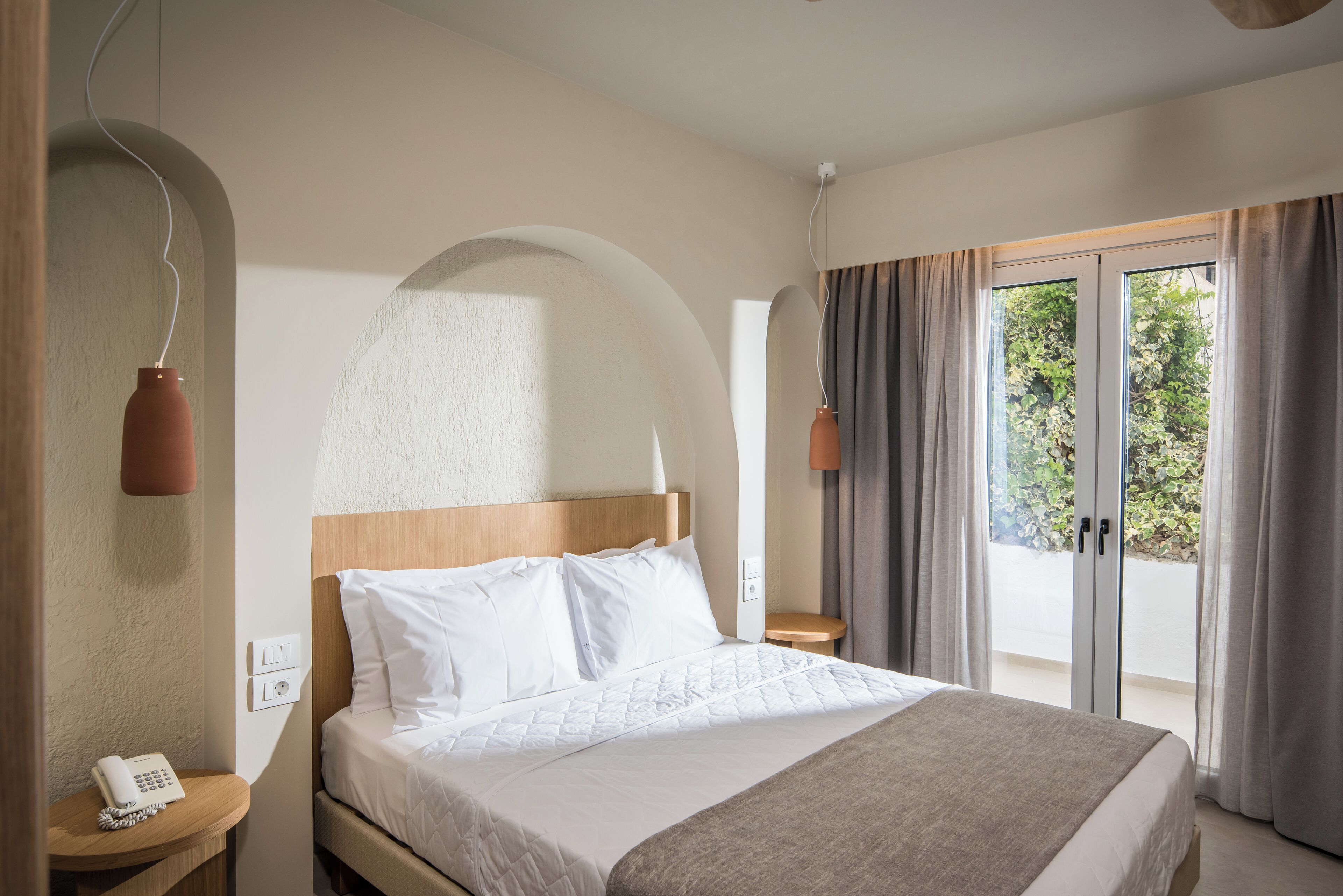 A hotel room with a bed, a bedside table with a phone, a window with curtains, and a view of greenery outside.