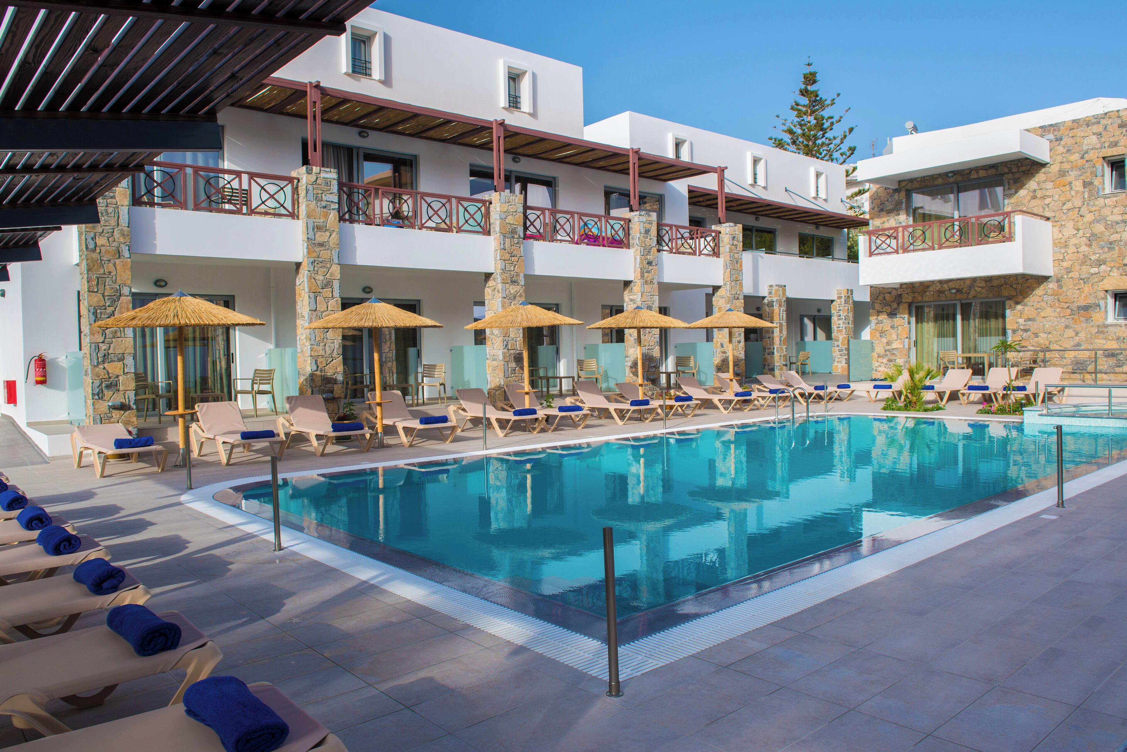Zona de piscina con sillones de descanso y sombrillas, una piscina azul cristalina y un edificio con balcones y pilares de piedra.