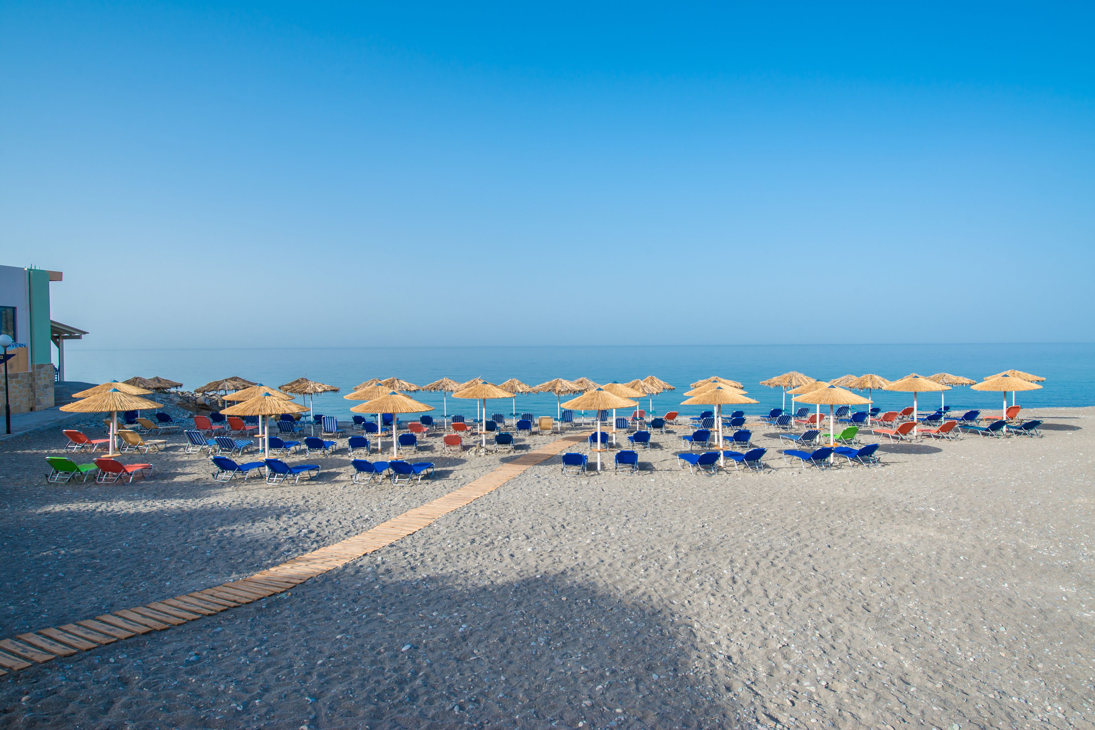 Een strand met rijen ligstoelen en parasols, een houten looppad en een gebouw aan de linkerkant.