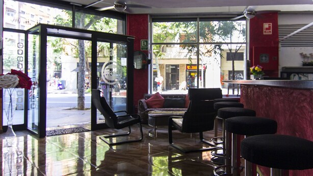 A modern interior with a glass door entrance, a seating area with chairs and a sofa, and a bar counter. - Go BCN Hi Sants - Hostel (Barcelona)