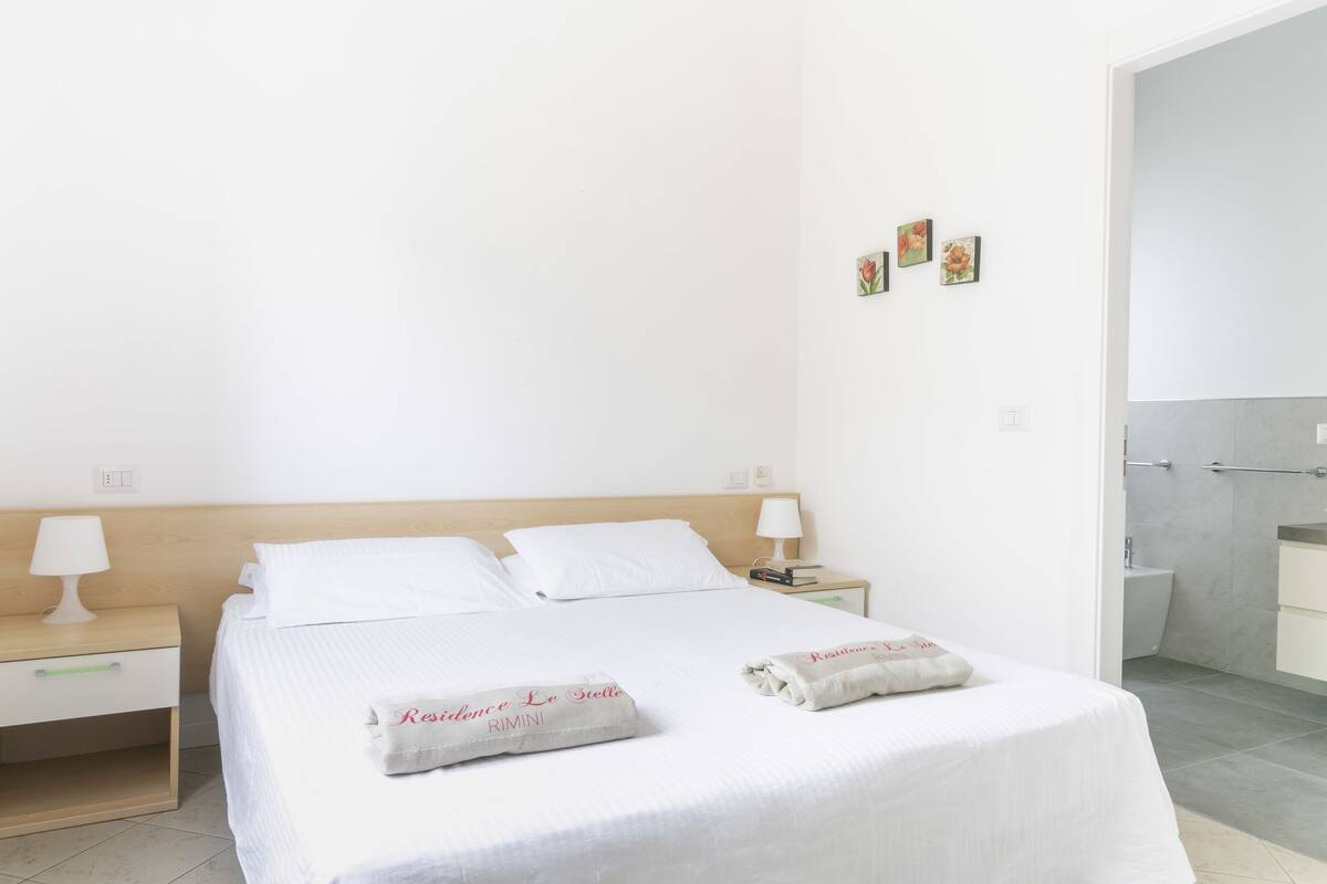A clean, modern hotel room with a large bed, bedside tables with lamps, a small bedside table with a book, and a bathroom visible through an open door.