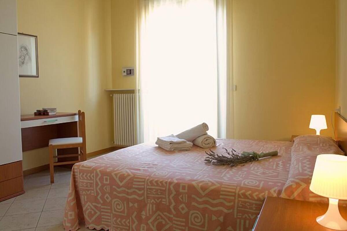 A hotel room with a bed, a desk, a chair, a radiator, a lamp, and a window with curtains.