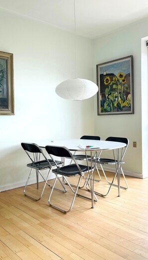 Un coin repas avec une table blanche, des chaises noires et un luminaire suspendu blanc. Un tableau représentant des tournesols est accroché au mur.
