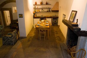 A cozy dining area with a wooden table set for four, surrounded by chairs. A fireplace is visible to the right, and a kitchen area with shelves and utensils can be seen in the background. - Palazzo Contino (Bagnoregio)