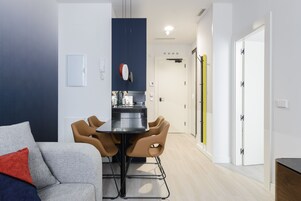 Ein modernes Wohnzimmer mit einem grauen Sofa, einem Esstisch mit vier Stühlen und einer Küchenzeile mit Kühlschrank und Herd.