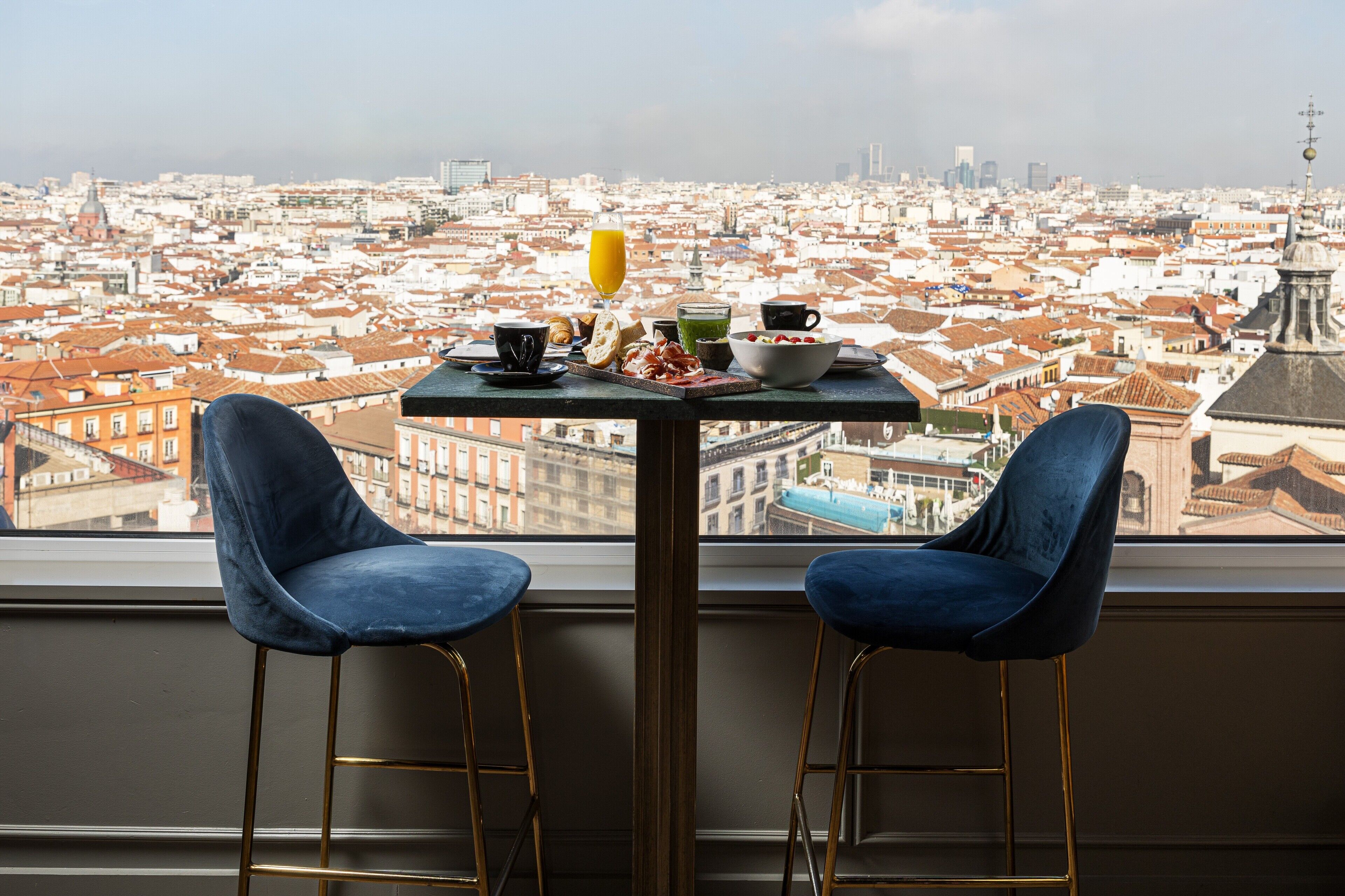 A table with breakfast items and two blue chairs in front of a window overlooking a cityscape.