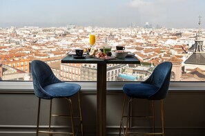 Una mesa con alimentos para el desayuno y dos sillas azules frente a una ventana con vistas a un paisaje urbano.