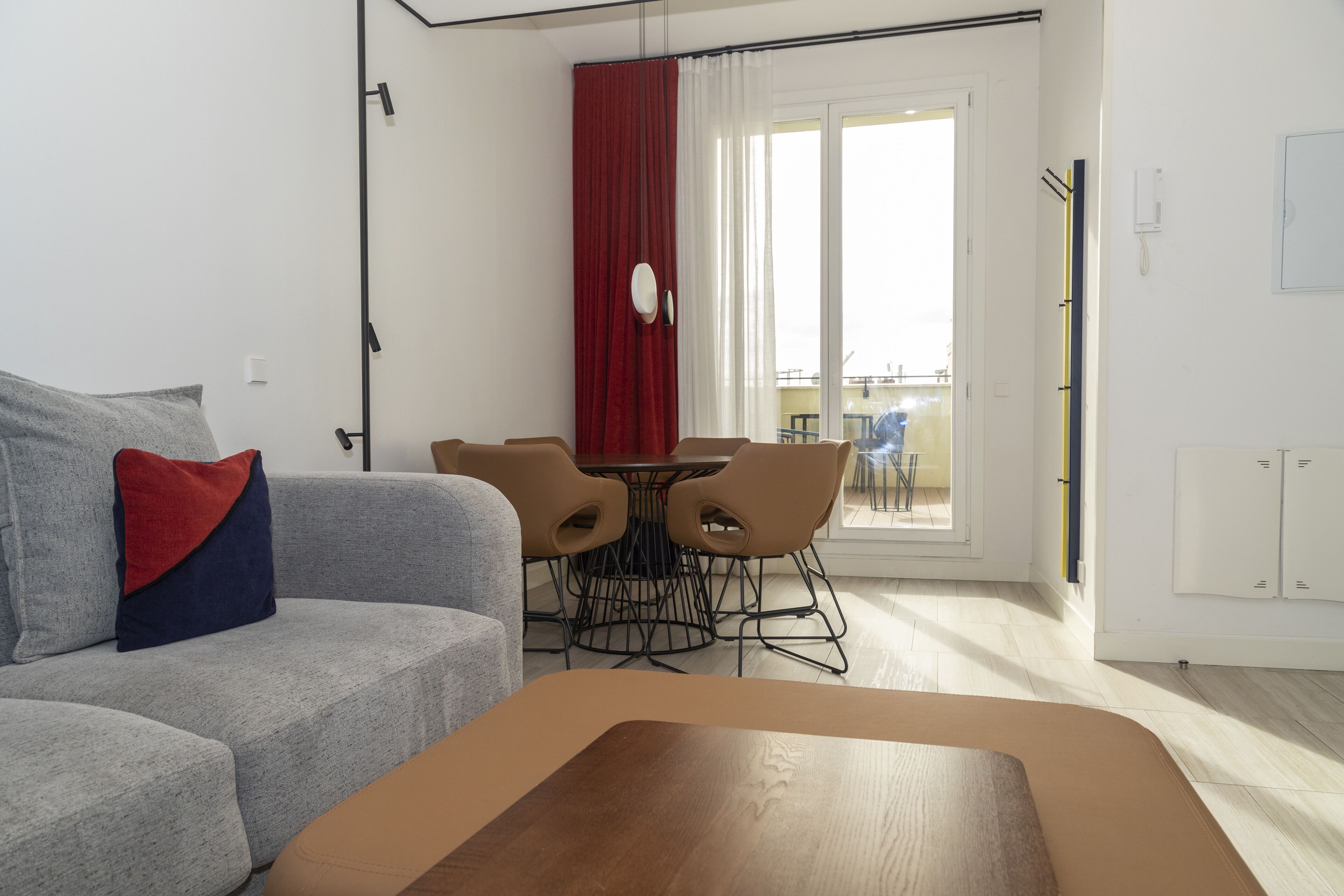 A modern living room with a grey sofa, a wooden coffee table, and a dining area with a red curtain and a small table.