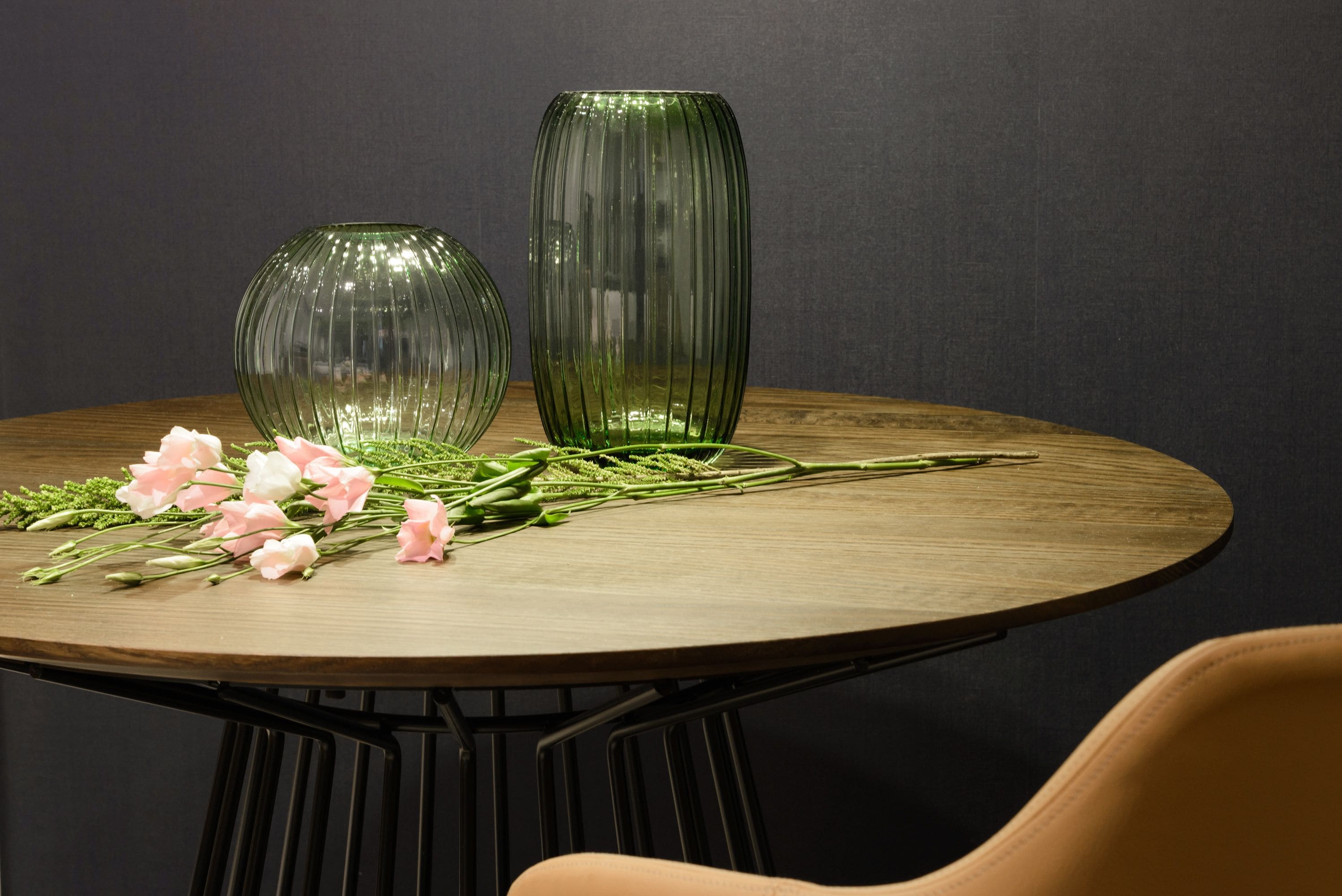 A round wooden table with two green glass vases and pink flowers on it.