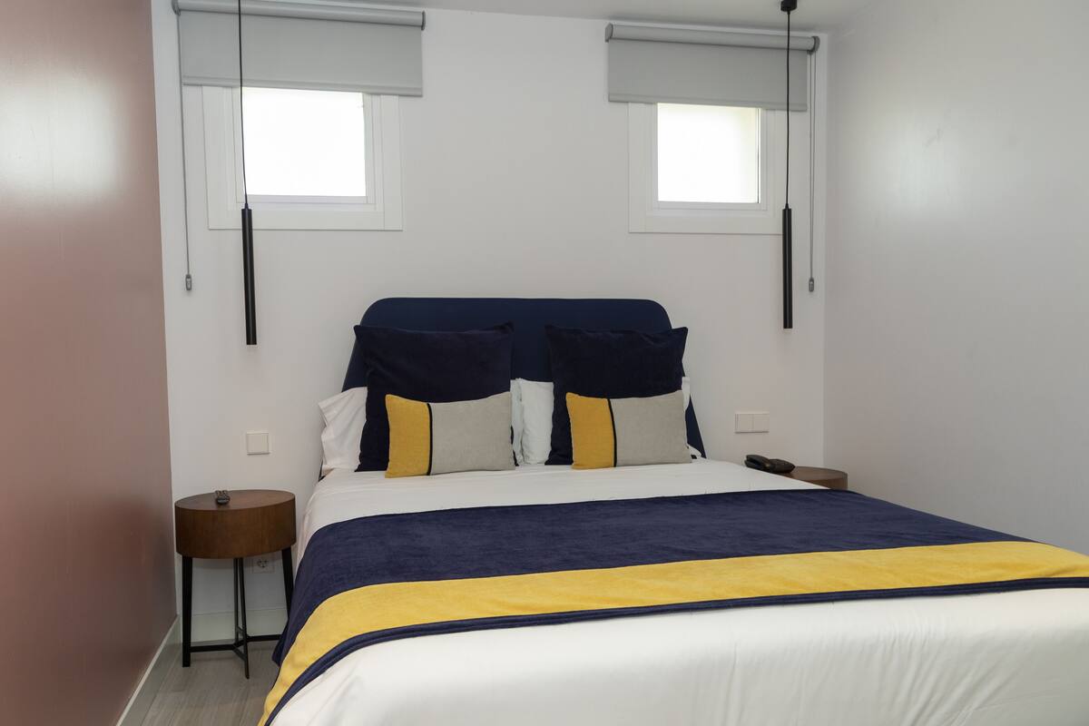 A hotel room with a bed, two bedside tables, a window with blinds, and a wall-mounted light fixture.