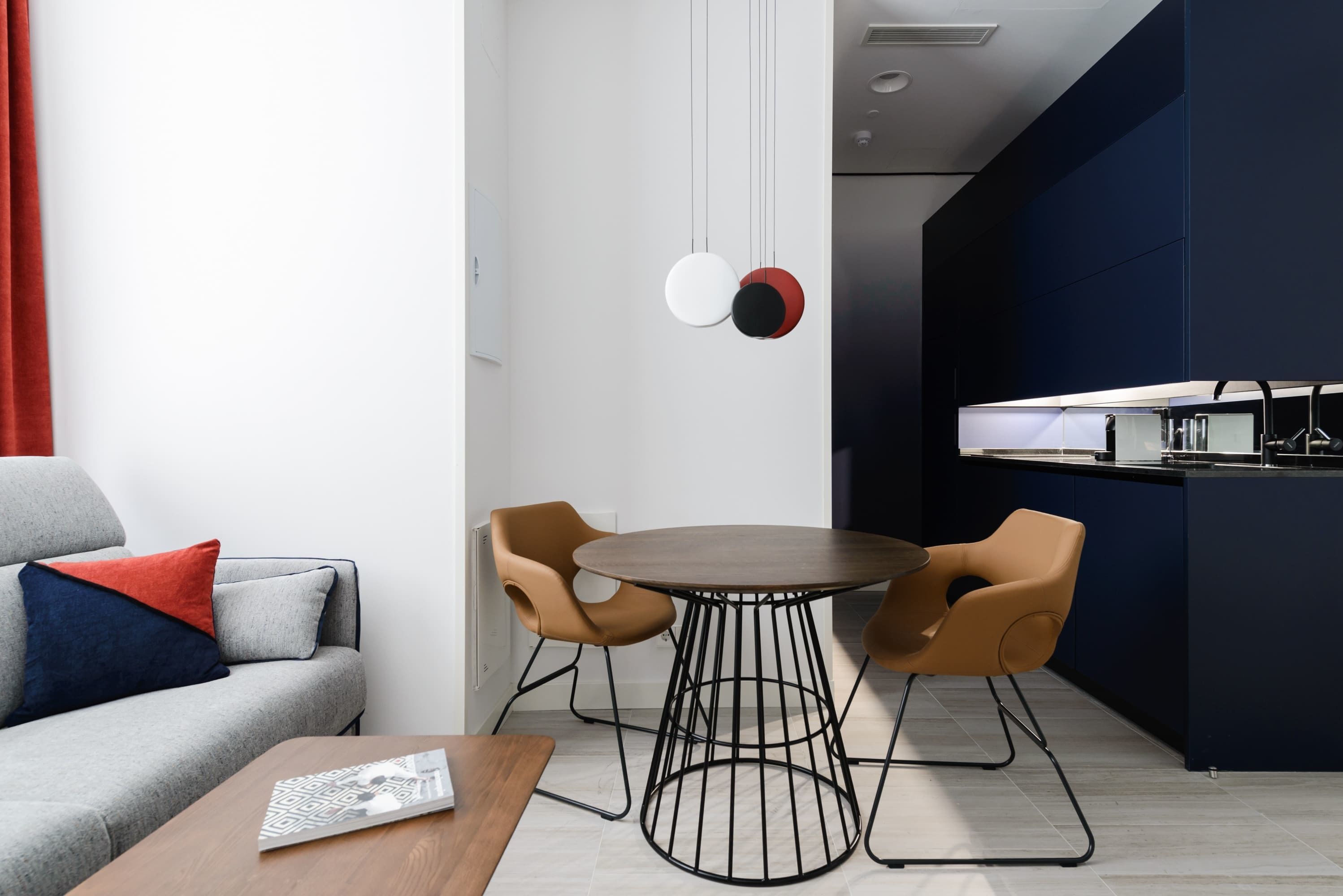Un salon moderne avec un canapé gris, deux fauteuils marron, une table ronde et un coin cuisine agrémenté d’un mur d’accent bleu foncé.