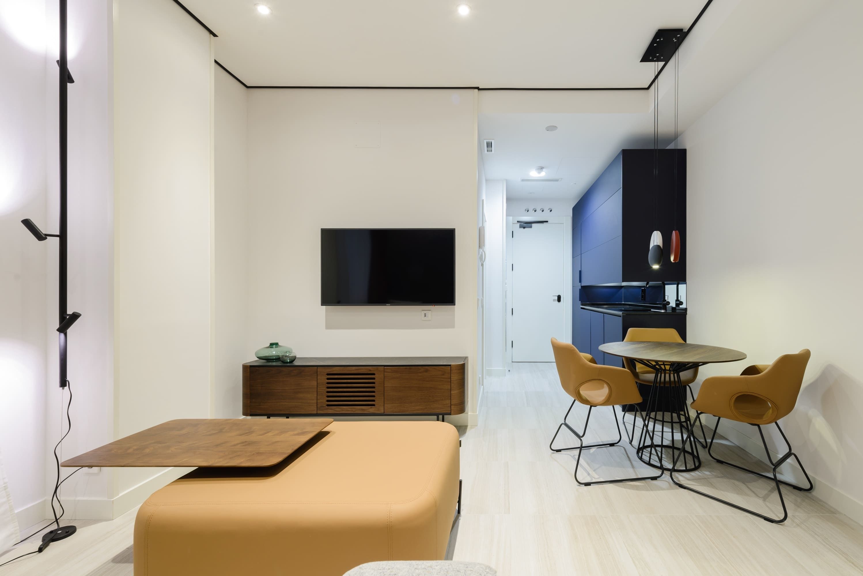 A modern, compact living space with a sofa, a dining table with chairs, a TV mounted on the wall, and a kitchenette in the background.