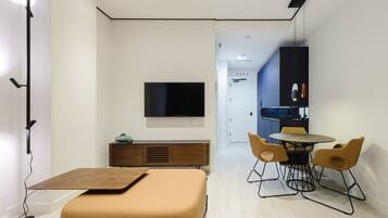 A modern, compact living space with a sofa, a dining table with chairs, a TV mounted on the wall, and a kitchenette in the background.