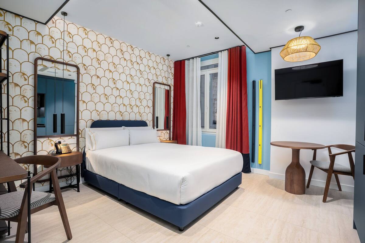 A modern hotel room with a large bed, a desk, a chair, a TV, and a window with curtains.