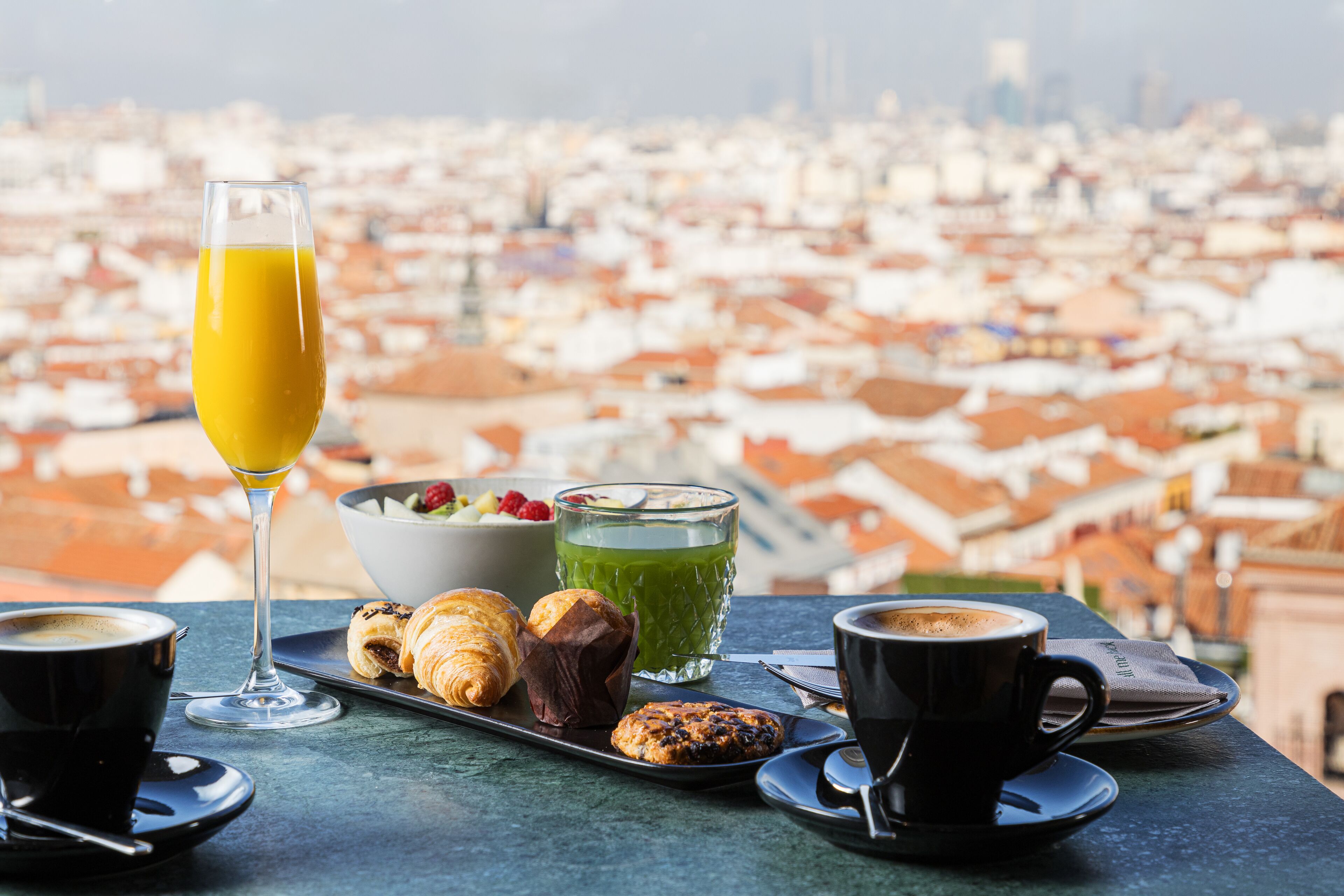 Une table dressée pour le petit-déjeuner, avec des croissants, une boisson verte et du café, le tout placé devant une fenêtre offrant une vue sur la ville.