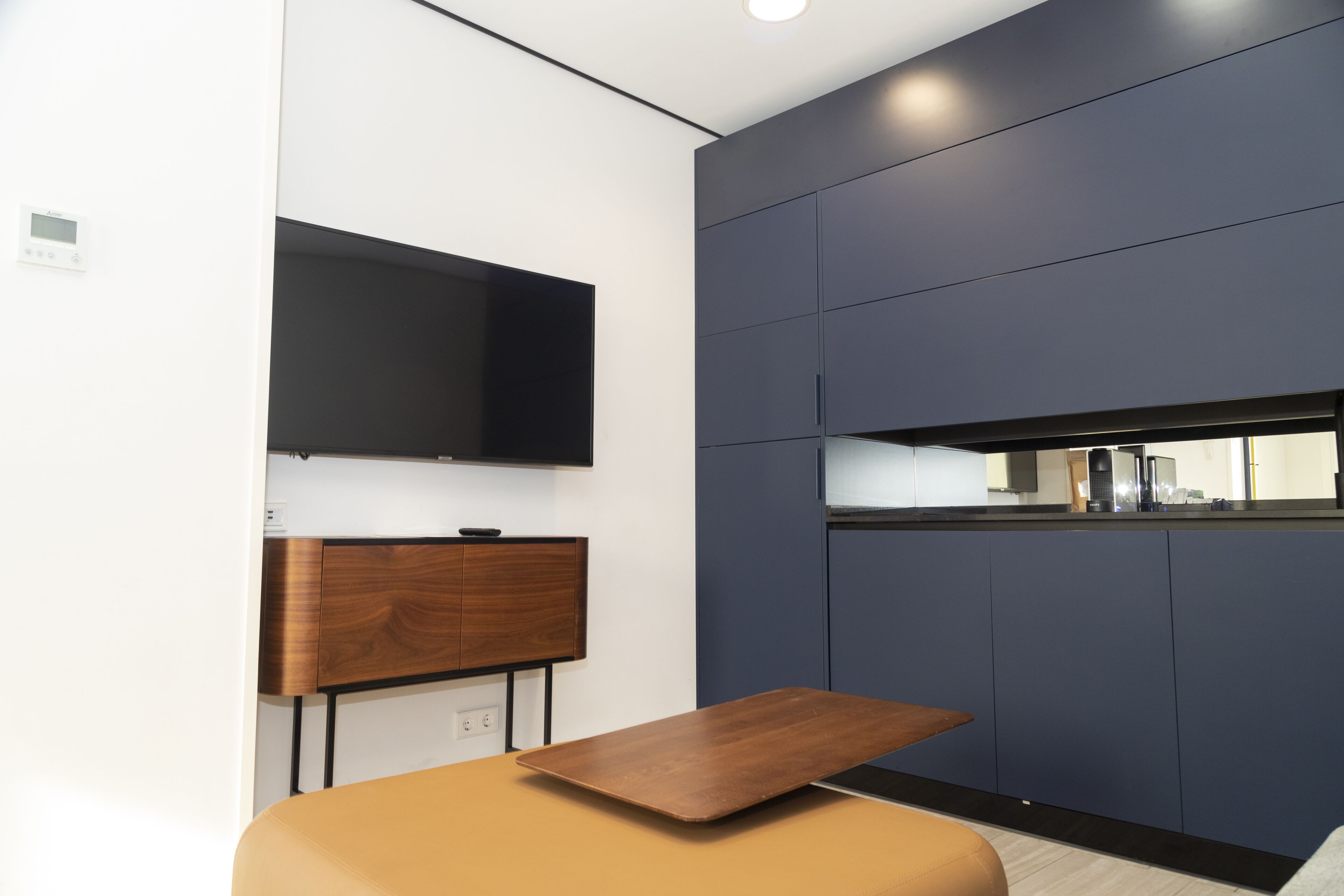 Een moderne keuken met een flatscreen televisie boven een houten kastje, een aanrecht met een koffieapparaat, en een klein tafeltje met een dienblad.