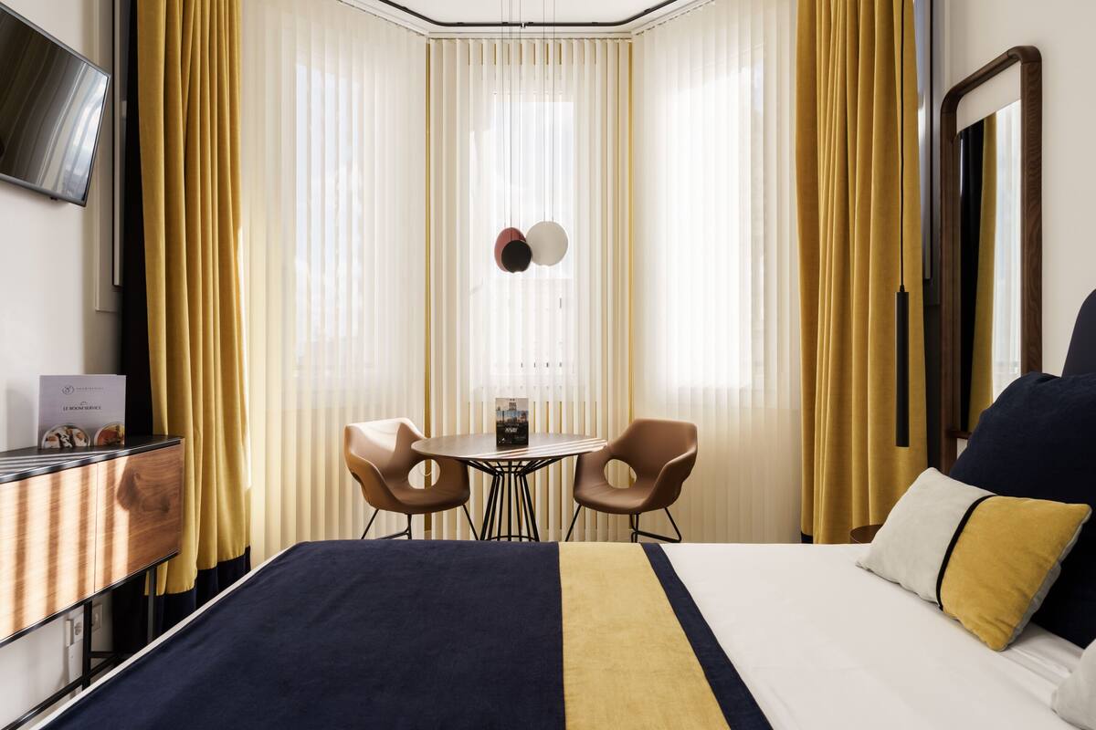 A hotel room with a bed, a desk, two chairs, a table, and a TV on the wall.