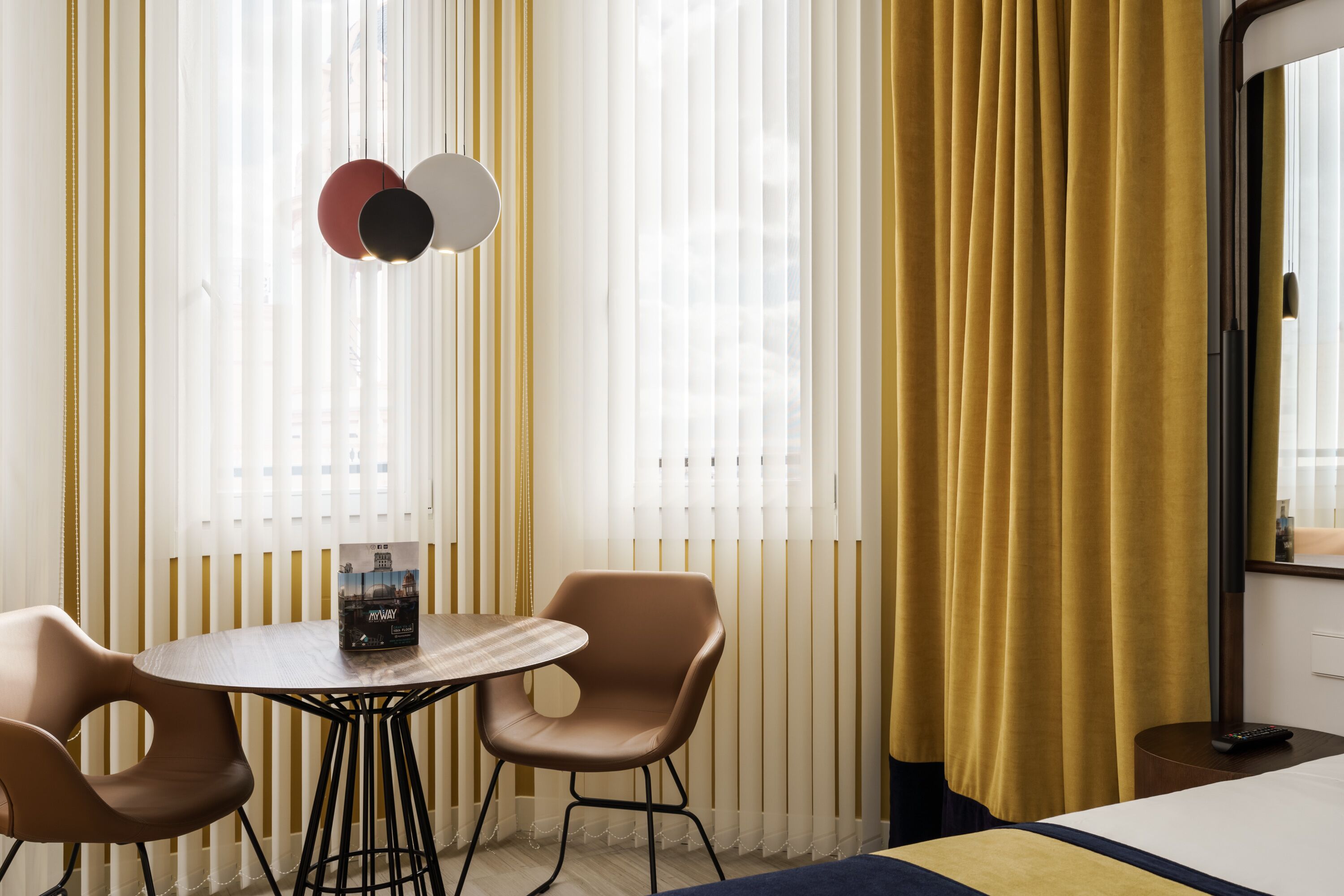 Une chambre d’hôtel avec une table ronde, deux chaises, un lit et une grande fenêtre avec des rideaux.