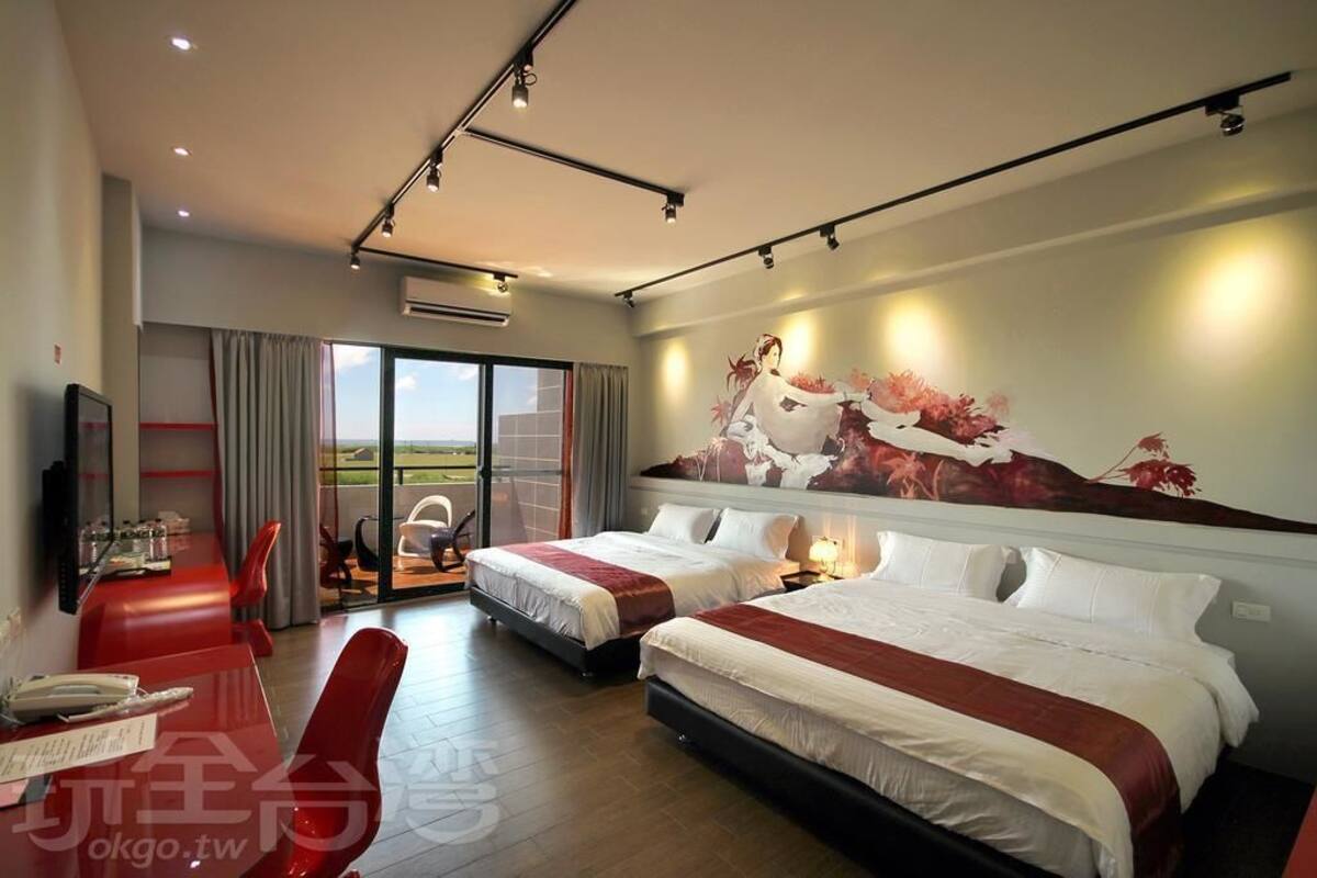 A hotel room with two beds, a dining area with red chairs, a large window with a view, and a painting on the wall.