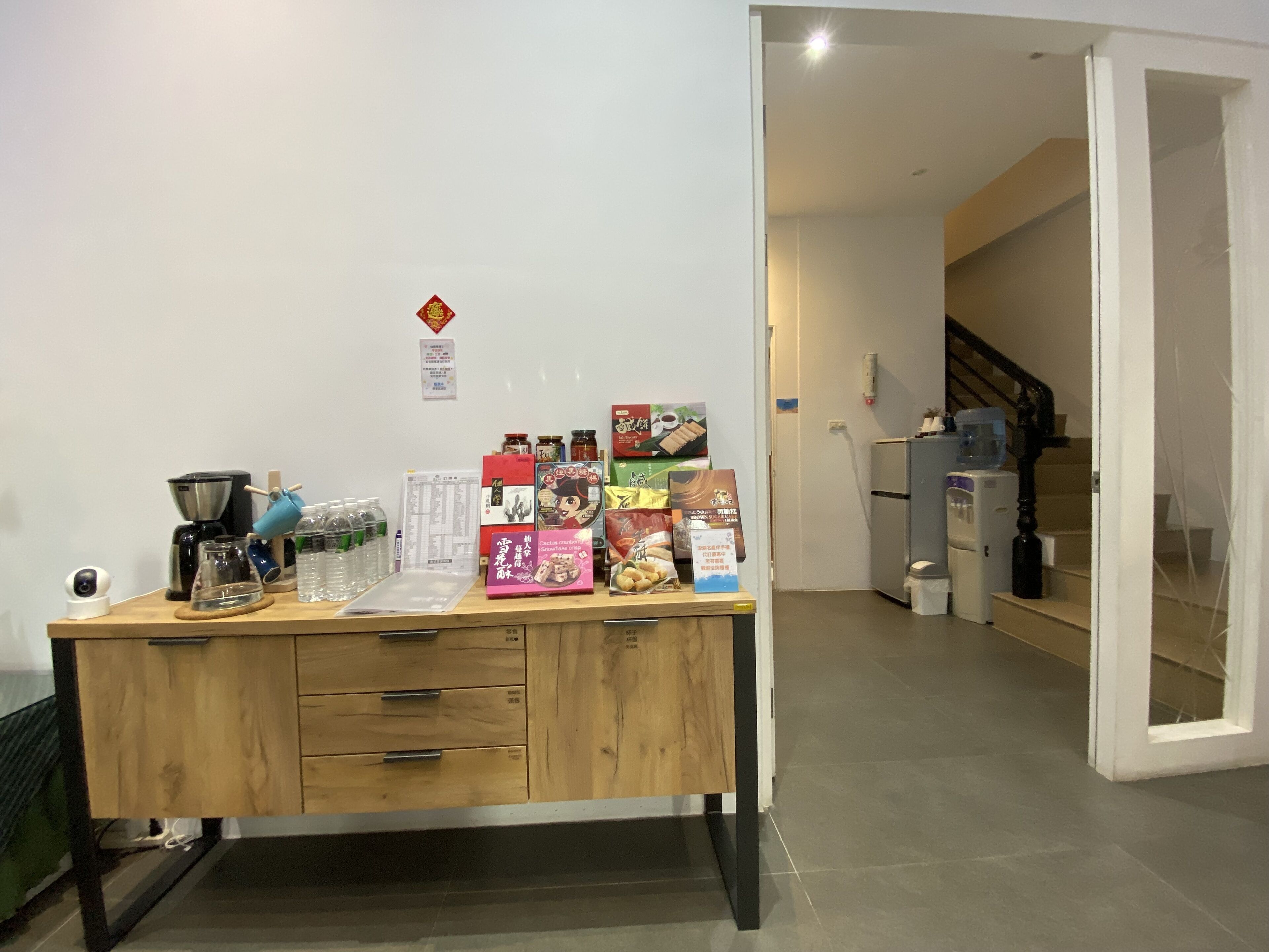 A wooden desk with various items including a coffee maker, glassware, and promotional materials. A camera is visible on the left side.