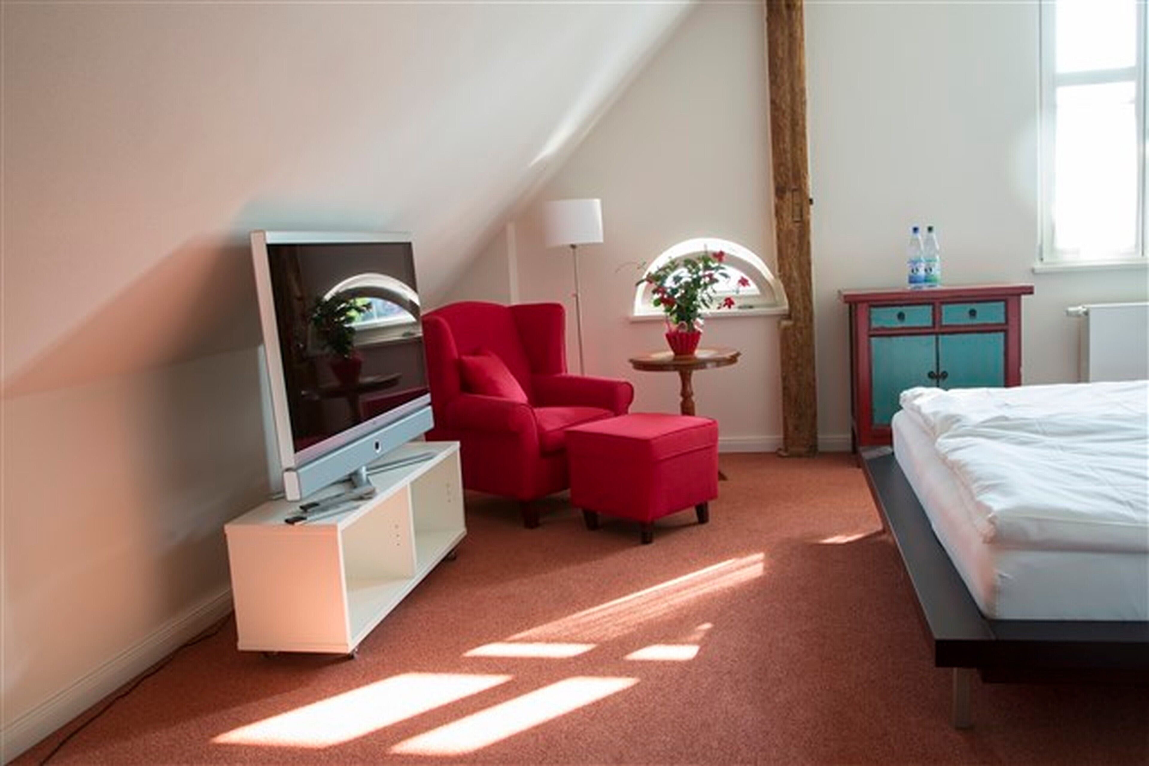A hotel room with a TV, red chair, ottoman, small table with a plant, and a bed.