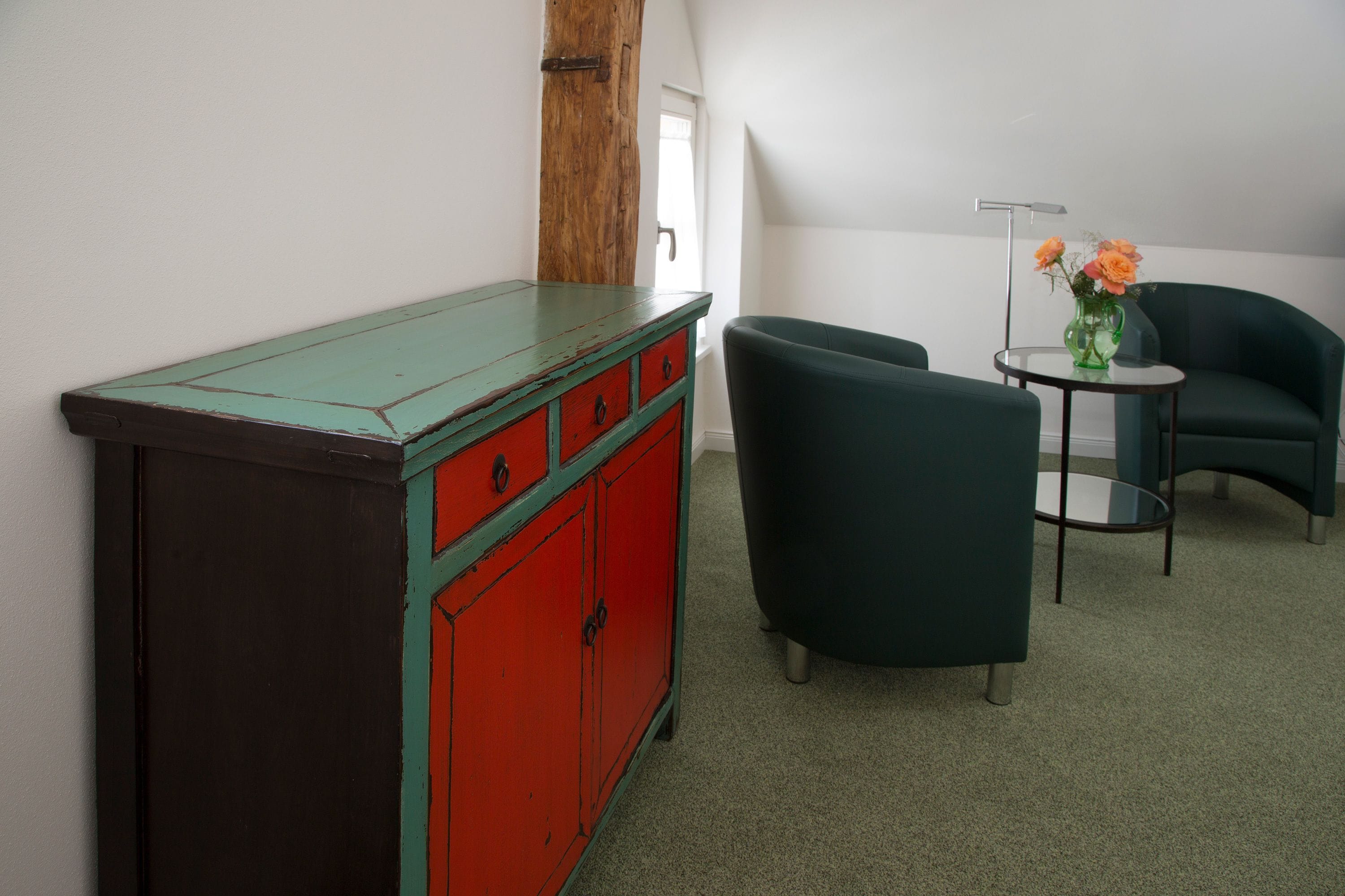 Een kamer met een groene fauteuil, een ladekast in rood en groen, en een klein tafeltje met een bloevas.