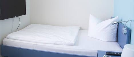Una cama individual con ropa de cama blanca, un televisor de pantalla plana montado en la pared, una mesita de noche con una lámpara y un control remoto.