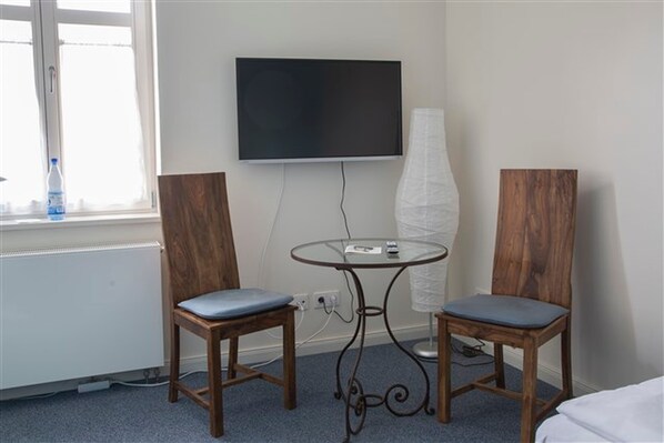 A hotel room with two wooden chairs, a glass table, a TV mounted on the wall, and a window with a water bottle on the sill.