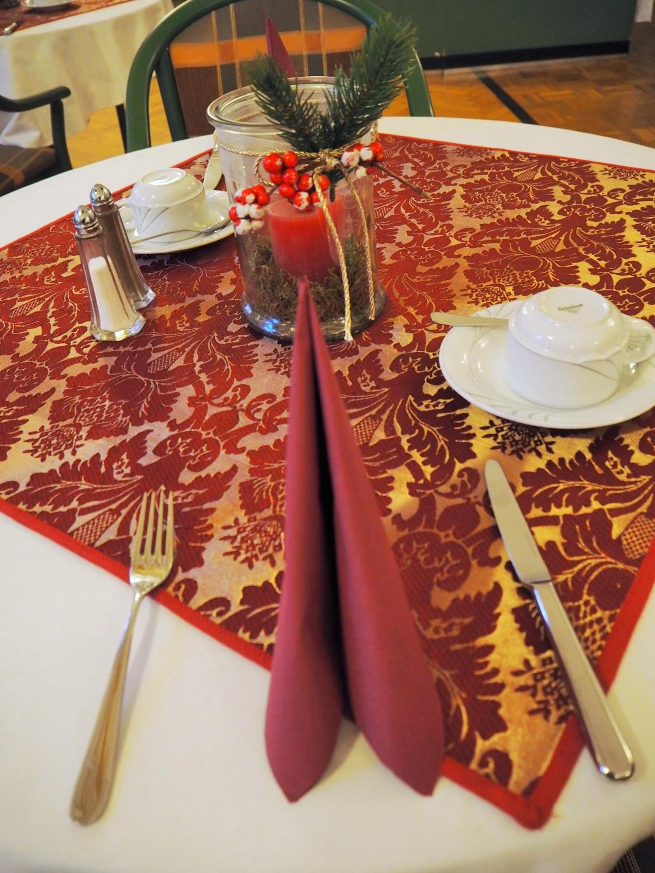 A table set with a white tablecloth, a red and gold patterned table runner, a glass candle holder with a pine branch and red berries, a white teapot, a white cup and saucer, a silver salt and pepper set, and a folded napkin.
