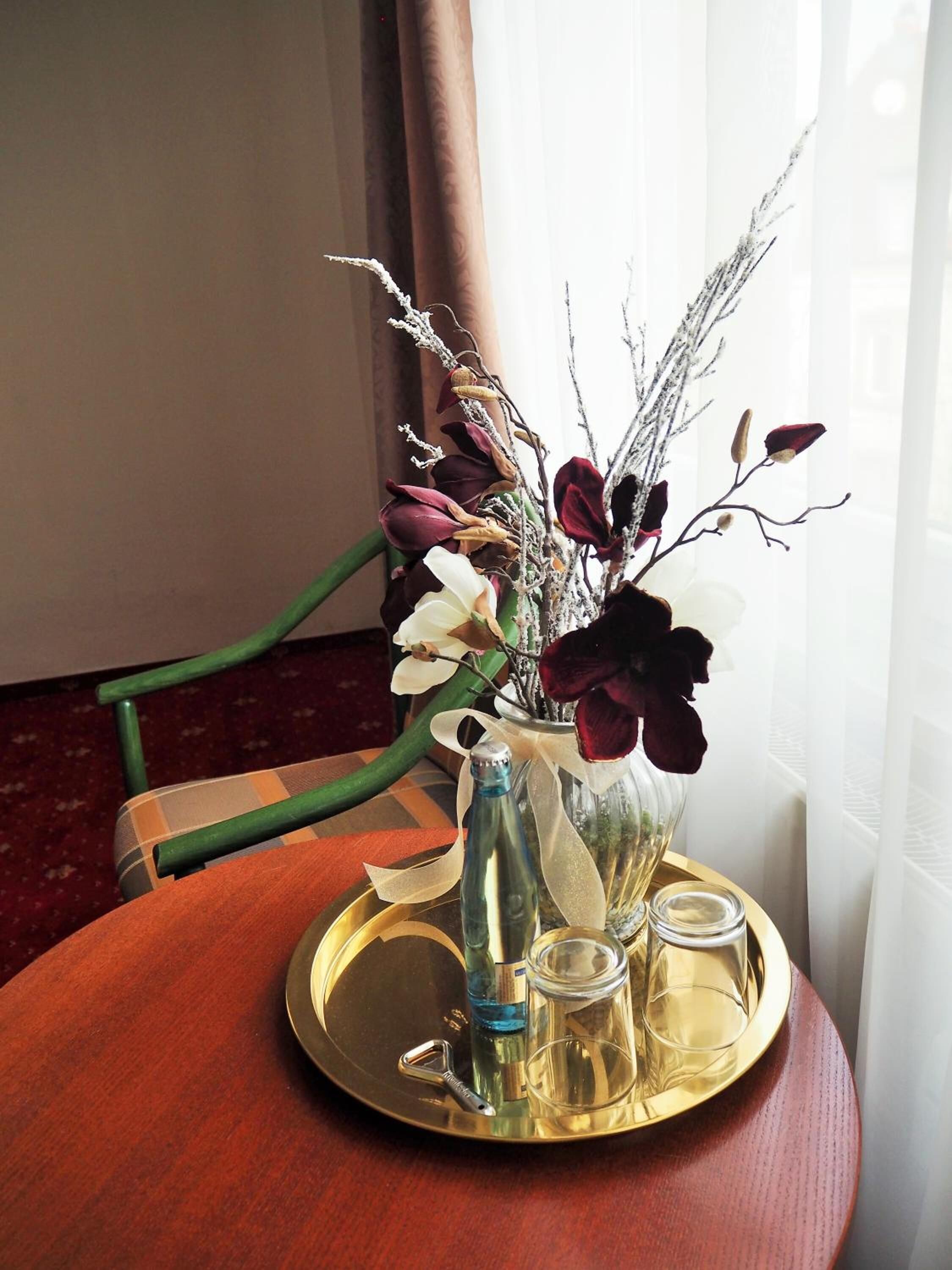 A round wooden table with a glass vase containing dried flowers and a metal bottle, accompanied by a gold tray with a glass and a metal bottle.
