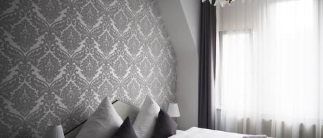 Une chambre d’hôtel moderne avec un papier peint à motifs, un lit recouvert de draps blancs, une lampe de chevet et une vue sur l’extérieur à travers des rideaux transparents.